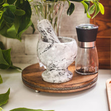 Load image into Gallery viewer, Marble mortar and pestle on a wooden stand with a pepper grinder and plant in the background.