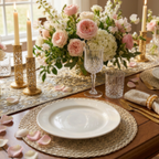 A styled dining table setting with a large floral centerpiece in pink and white roses, gold candlesticks, clear textured glassware, woven placemats, and white plates on a wooden table.