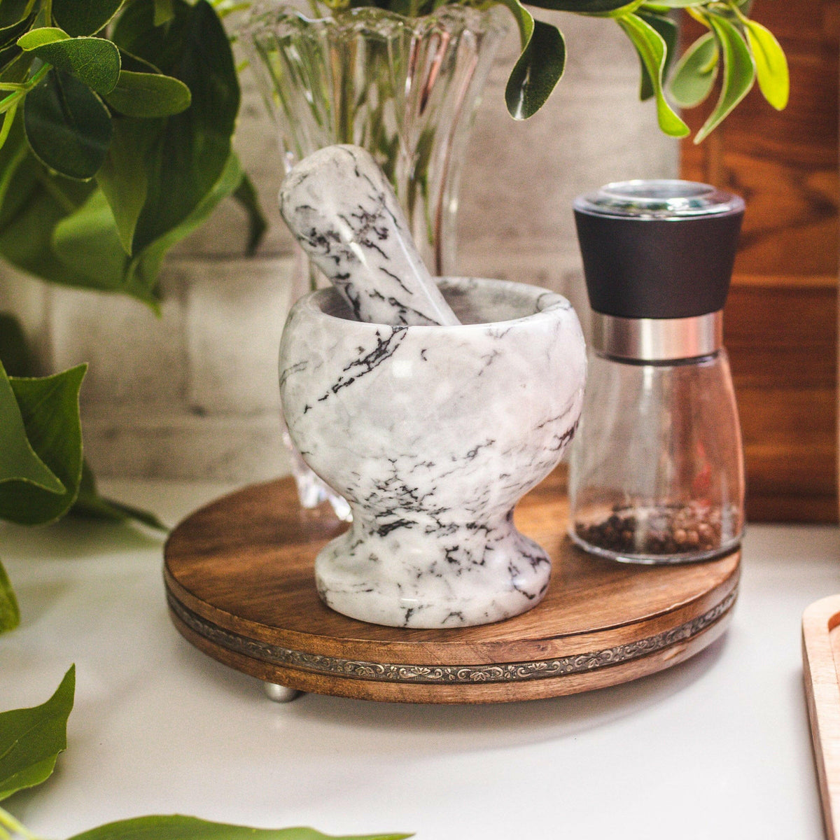 Marble mortar and pestle on a wooden stand with a pepper grinder and plant in the background.